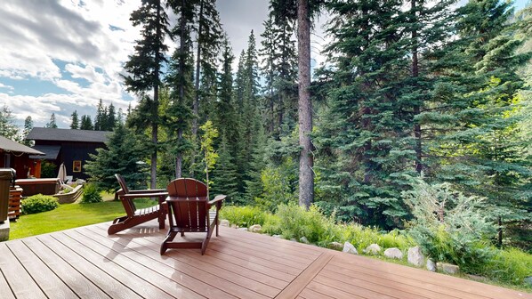 Une terrasse en bois, agrémentée de mobilier d’extérieur, entourée d’une forêt dense et avec une maison en arrière-plan.