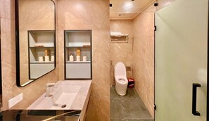 Egy modern fürőszoba, márvány mosdókagylóval, két polccal, melyeken piperecikkek találhatók, egy WC-vel és egy üvegbetéttel ellátott zuhanyzóval.