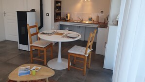 A dining area with a round table, two wooden chairs, a small table with a magazine and a cup, and a kitchen in the background. - Gîte PMR Balade Océane Lodonnec (Loctudy)