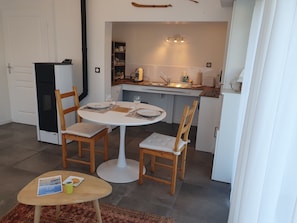Un comedor con una mesa redonda, dos sillas de madera, una mesita con una revista y una taza, y una cocina al fondo.