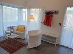 A living room with a white sofa, a small table with a lamp, a rug, and a wall-mounted coat rack with a jacket. - Gîte PMR Balade Océane Lodonnec (Loctudy)