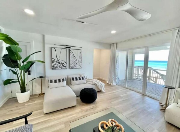 A modern living room with a large potted plant, two sofas, a coffee table, and a view of the ocean through large sliding doors.