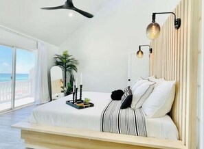A modern bedroom with a large bed, a ceiling fan, a view of the ocean, and a potted plant.