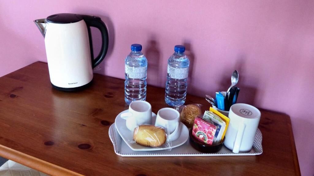 Una mesa de madera con una bandeja que contiene una cafetera blanca, dos botellas de agua, un plato con un pastel, una taza, una bolsita de té y una cuchara.