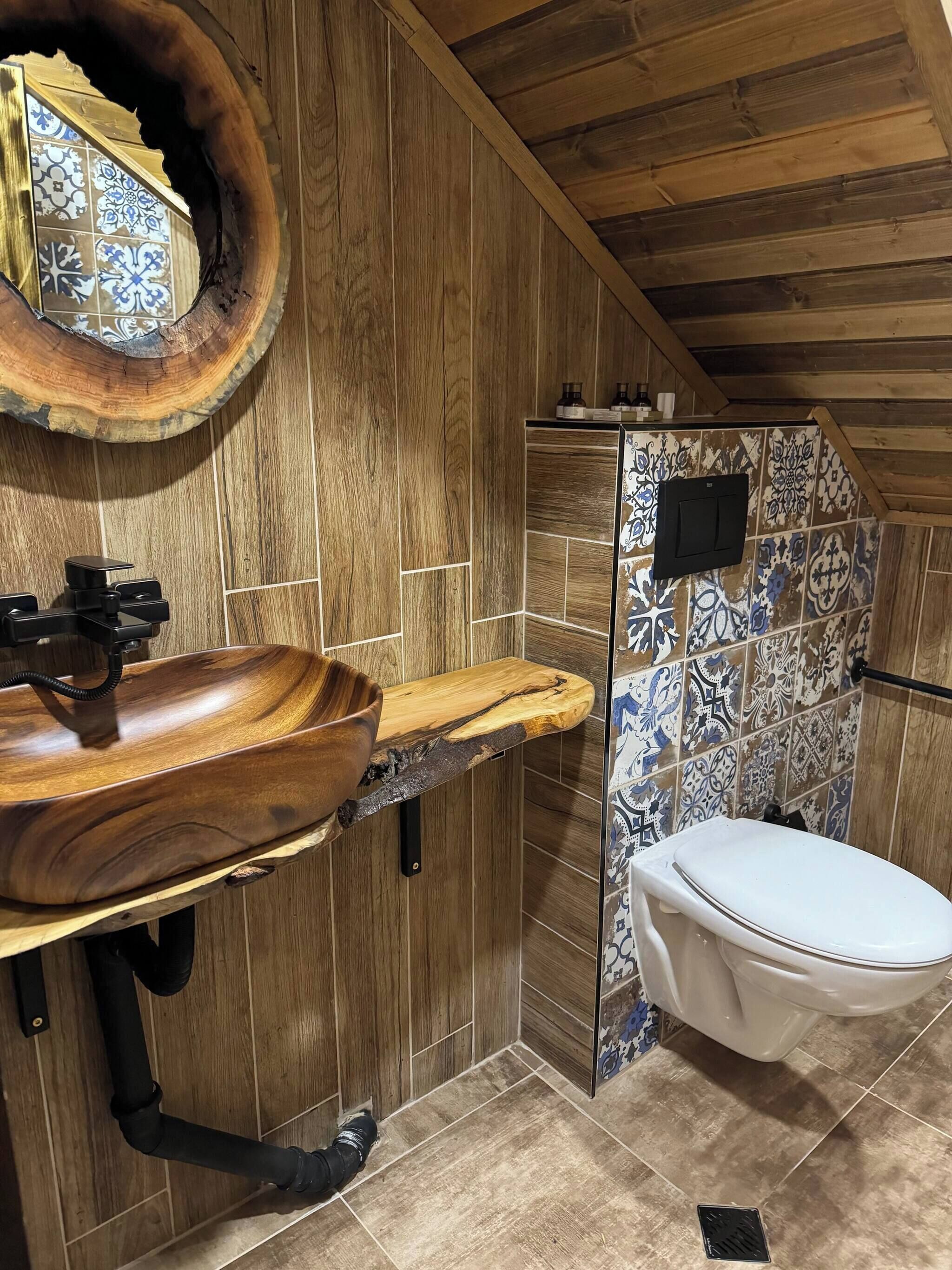 Un baño con tocador de madera, lavamanos, inodoro y zona de ducha con azulejos estampados.