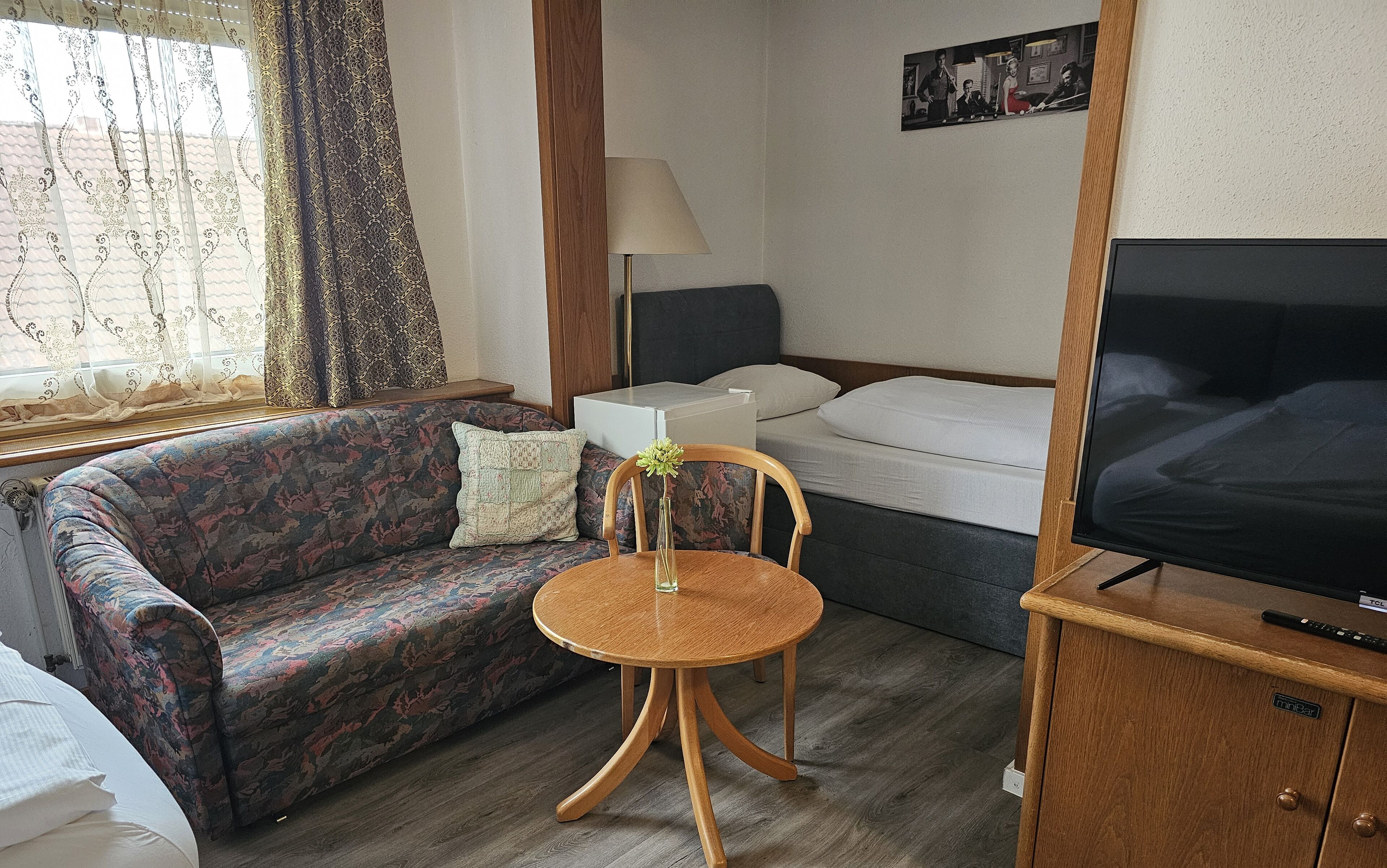 Camera d'albergo compatta con un divano a motivi floreali, un piccolo tavolino rotondo in legno con un vaso, un letto con biancheria bianca, una televisione su un mobile in legno e una lampada.