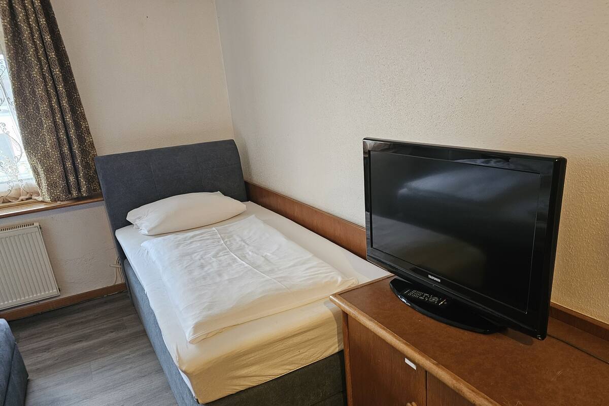 Una cama individual con colchón blanco y cabecera gris, un televisor sobre un mueble de madera, y una ventana con cortinas estampadas.