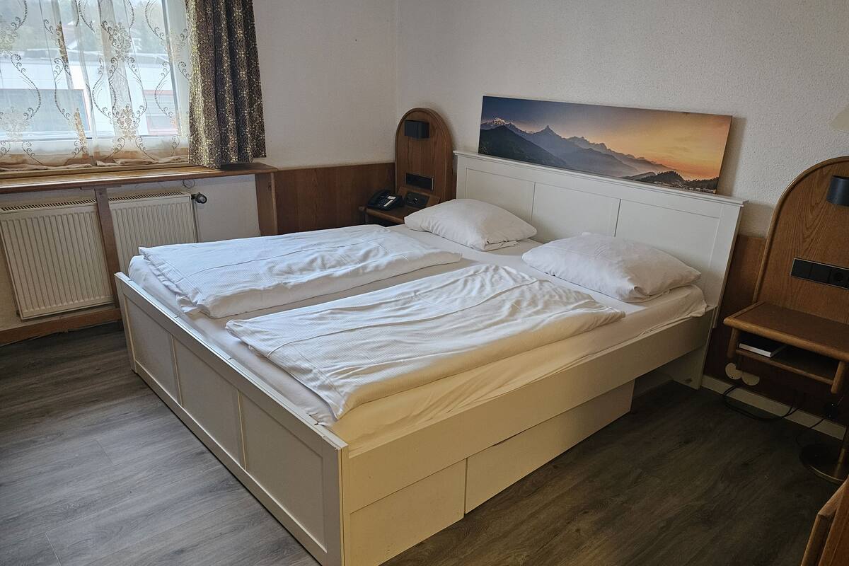 Una cama doble con sábanas blancas, un cabecero de madera, una mesita de noche con lámpara y un cuadro de un paisaje montañoso en la pared.