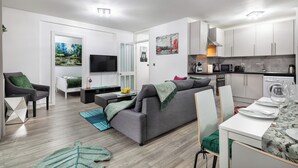 A modern living room with a grey sofa, a dining table with chairs, a TV, and a kitchen area with cabinets and appliances.