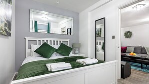 A modern bedroom with a white bed, green pillows, a green bedspread, and a white nightstand with a lamp.
