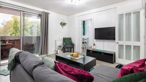 A modern living room with a sofa, a coffee table with fruit, a TV, and a sliding glass door leading to an outdoor area with a table and chairs.