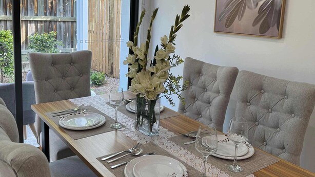 A dining area with a wooden table set for four, upholstered chairs, and a vase with flowers. - Stylish Family 3BR  in Central Hope Island, Family (Hope Island)