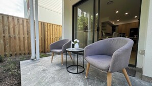 An outdoor patio with two grey armchairs and a small round table, a wooden fence, and a glass door leading to a kitchen area. - Stylish Family 3BR  in Central Hope Island, Family (Hope Island)