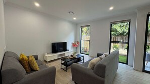 A modern living room with two grey sofas, a flat-screen TV, a coffee table, and a large window offering a view of a backyard. - Stylish Family 3BR  in Central Hope Island, Family (Hope Island)