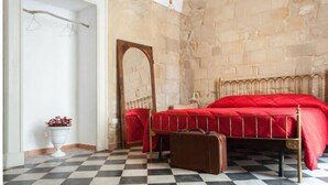 Ein Zimmer mit Schachbrettmuster-Boden, einem Bett mit roter Tagesdecke, einem Holzkopfende, einer weißen Vase mit roten Blumen und einer Steinstur.