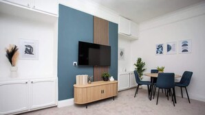 Un espace repas moderne avec un téléviseur fixé sur un mur bleu, un meuble en bois et une table ronde entourée de chaises.