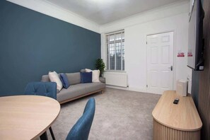 A room with a grey sofa, a round table, a wooden counter, a radiator, a door, and a window with blinds.