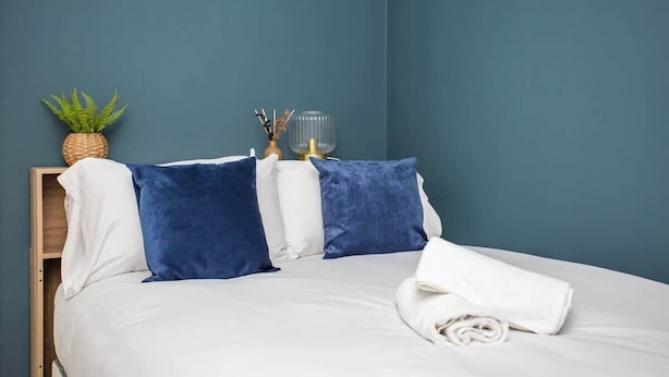 Un lit avec une literie blanche, des oreillers bleus, une serviette blanche enroulée, une petite plante en pot et une lampe en verre posée sur une table de chevet en bois.