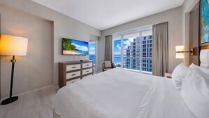 Ein Hotelzimmer mit einem großen Bett, einem an der Wand befestigten Fernseher, einem Schreibtisch mit Stuhl und Blick auf die Stadt durch große Fenster.