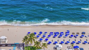 A beach with rows of blue umbrellas and lounge chairs, a lifeguard tower, and palm trees. - Holiday Waves: Oceanview Charm at Central Beach (Fort Lauderdale)