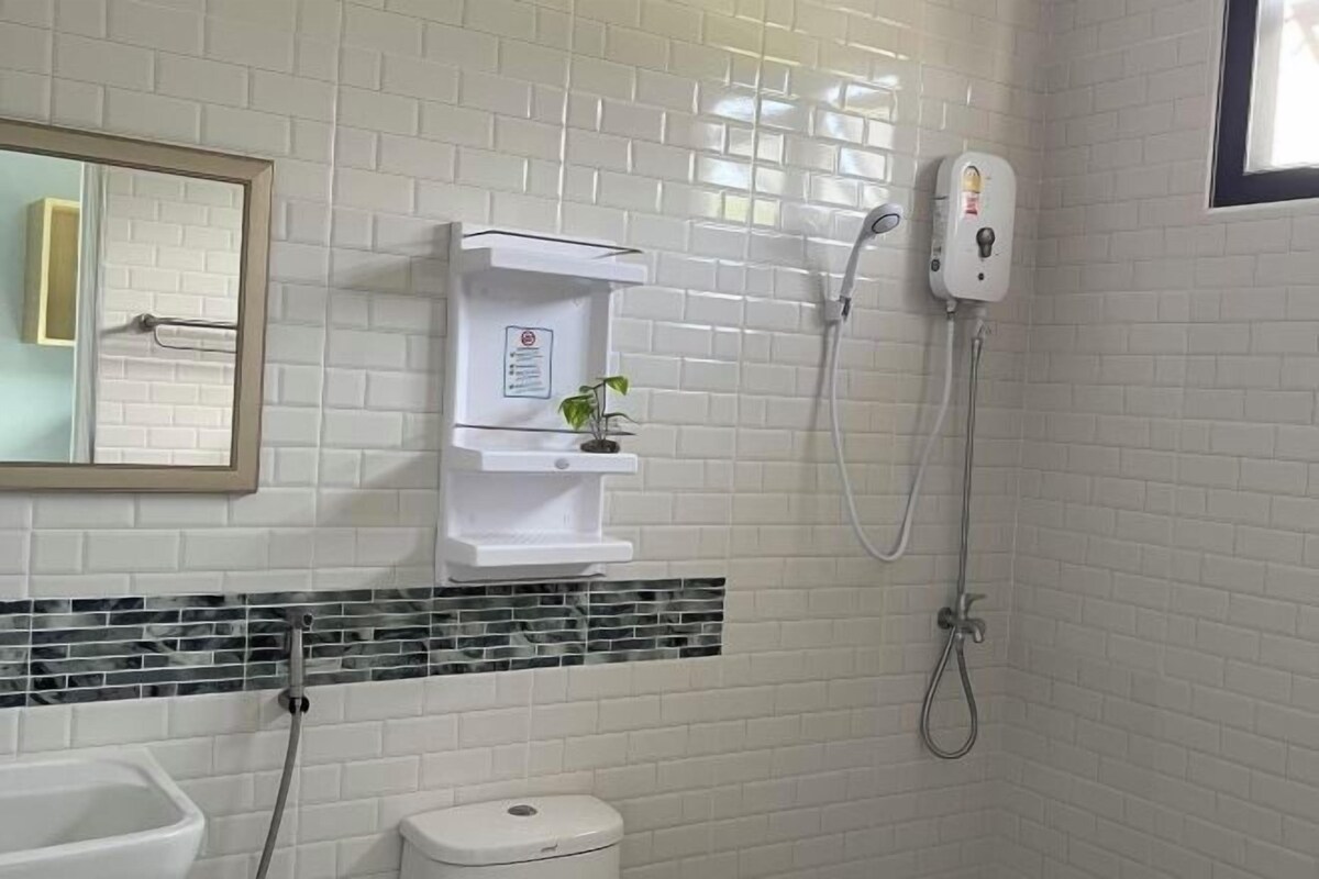 Un baño con paredes de azulejos blancos, un lavamanos blanco, un inodoro y una ducha con cabezal de ducha manual.