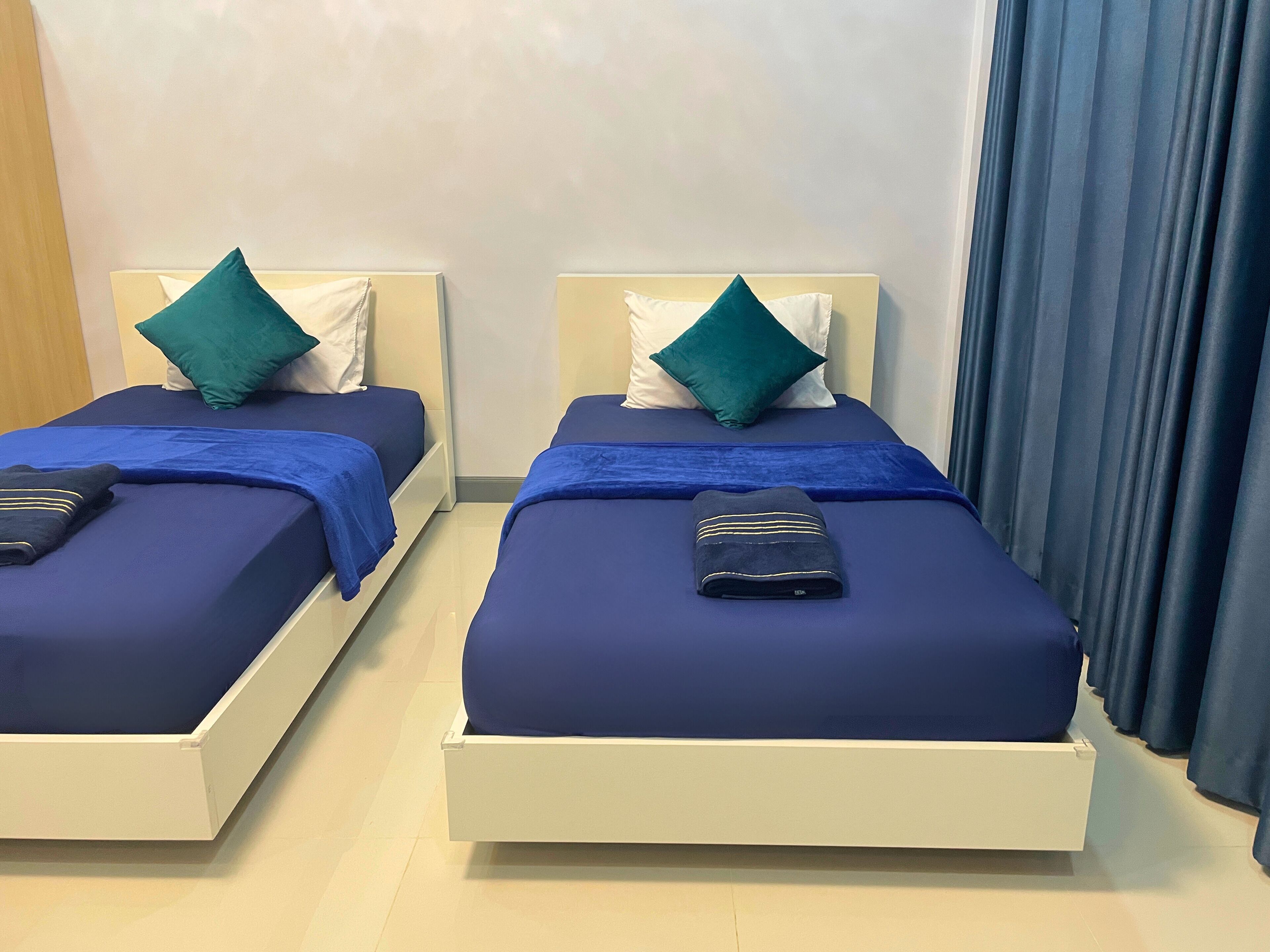 Zwei Einzelbetten mit blauer Bettwäsche und grünen Kissen, ein gefaltetes Handtuch auf jedem Bett und ein Fenster mit Vorhängen.