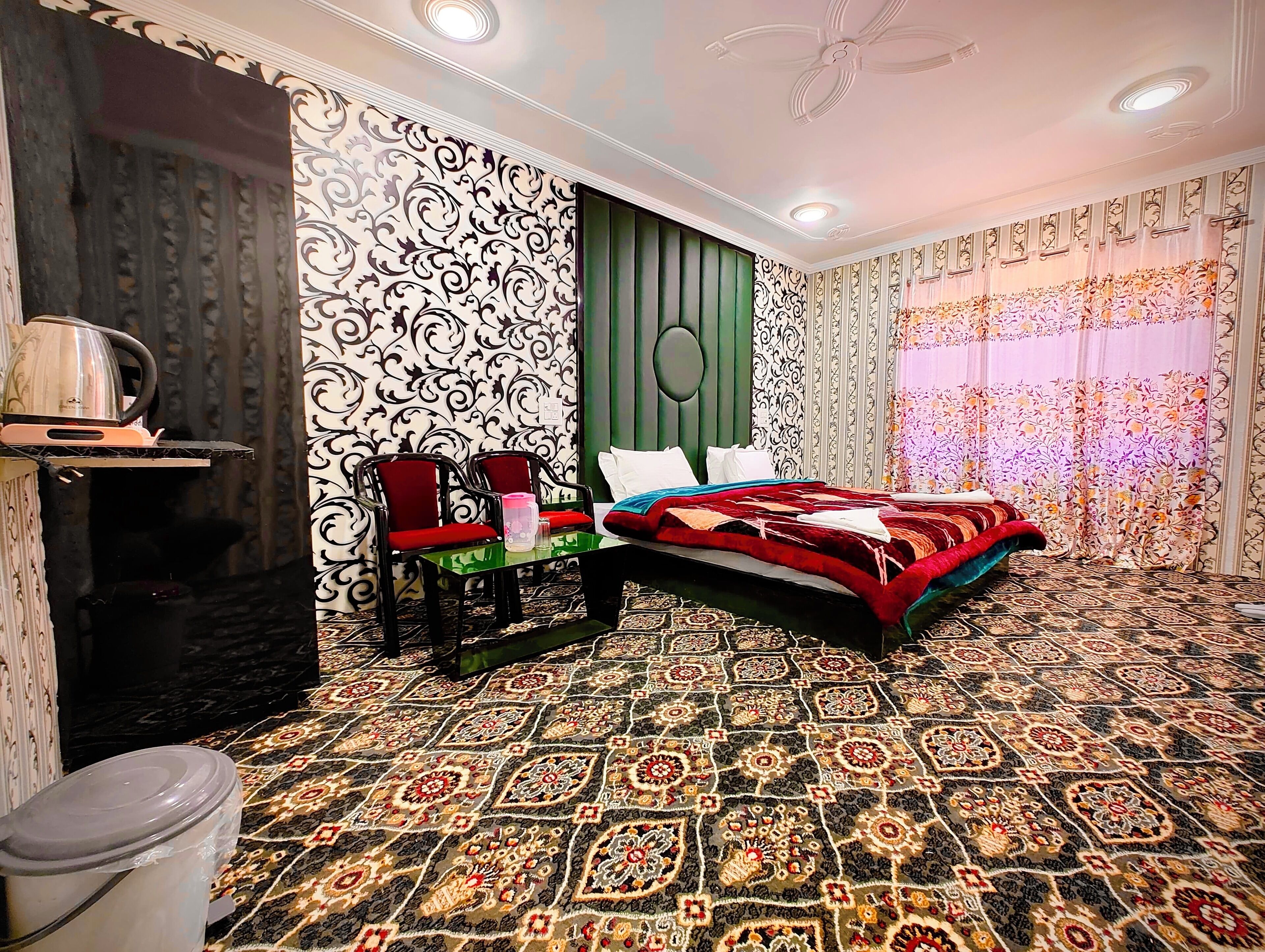 A bedroom with a patterned carpet, a bed with red bedding, a green accent wall, and a decorative ceiling fan.