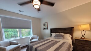 Une chambre à coucher comprenant un lit, deux chaises, une table de chevet, une lampe, un ventilateur de plafond, une fenêtre avec des stores et un tableau accroché au mur.