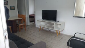 A living room with a TV on a white stand, a sofa, a dining table with chairs, and a door leading to another room. - Round trip - LA18-Bertioga, SESC, Riviera, Vista Linda, Indaiá (Jardim Rafael Bertioga)