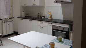 Eine kleine Küche mit weißen Schränken, einer schwarzen Arbeitsplatte und einem Tisch mit gemustertem Tischläufer. Bunte Flaschen stehen auf den Schränken und auf dem Tisch steht eine Kaffeetasse.