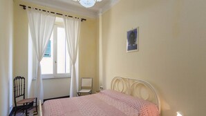 Ein Schlafzimmer mit einem Bett, einem Sessel, einem Nachttisch, einem Fenster mit Vorhängen und einem an der Wand hängenden Bild.