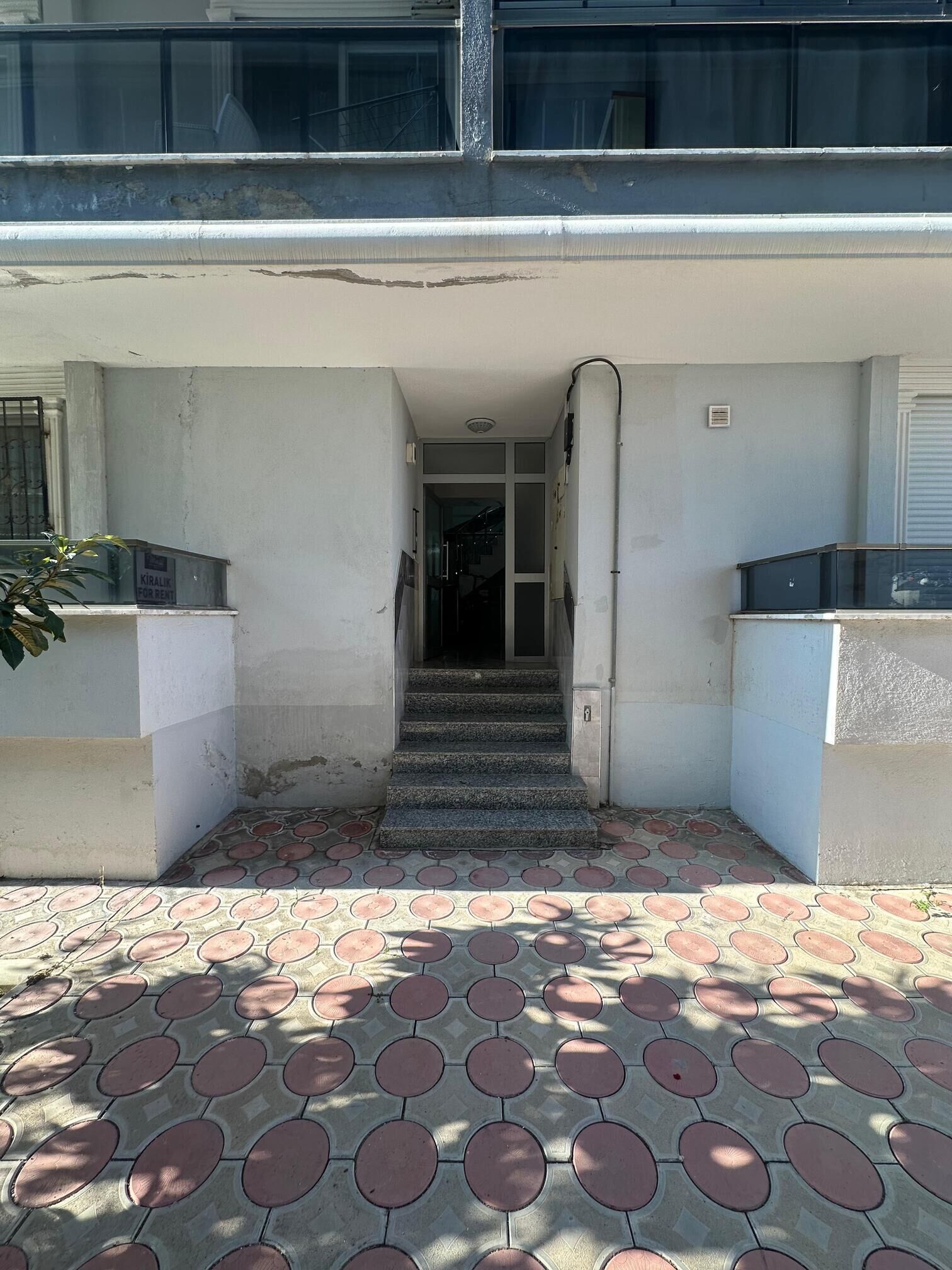A building entrance with a tiled pavement, a staircase leading to an interior door, and a sign indicating availability for rent.