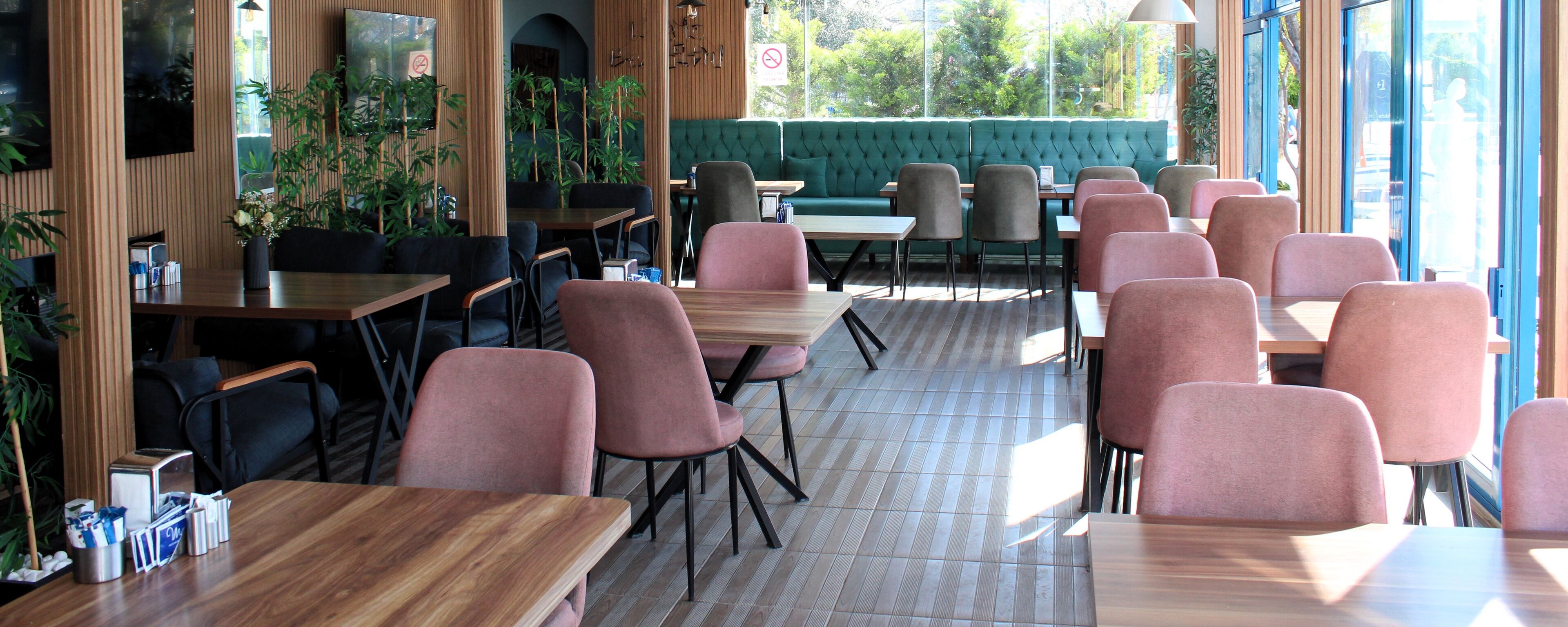 Un restaurante con mesas y sillas de madera, un sofá verde y ventanas amplias.