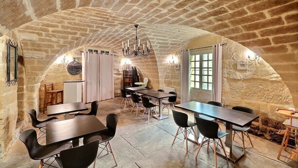 Une pièce rustique aux murs de pierre, comprenant un coin repas avec des tables en bois et des chaises noires, un coin cuisine avec un réfrigérateur et un plan de travail, et une grande porte en arc.