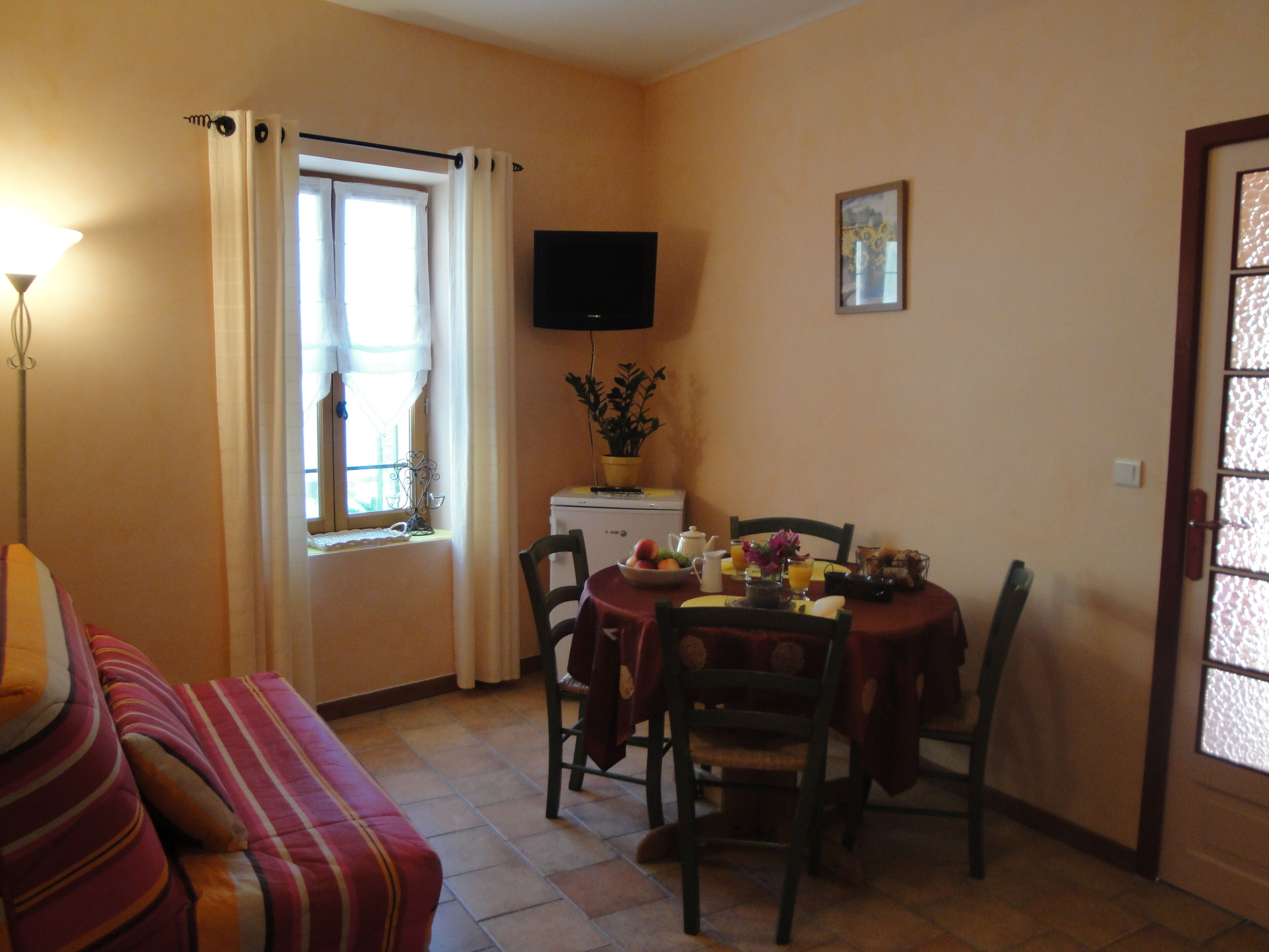 A cozy living room with a sofa, a dining table set for two, a flat-screen TV mounted on the wall, and a window with curtains.
