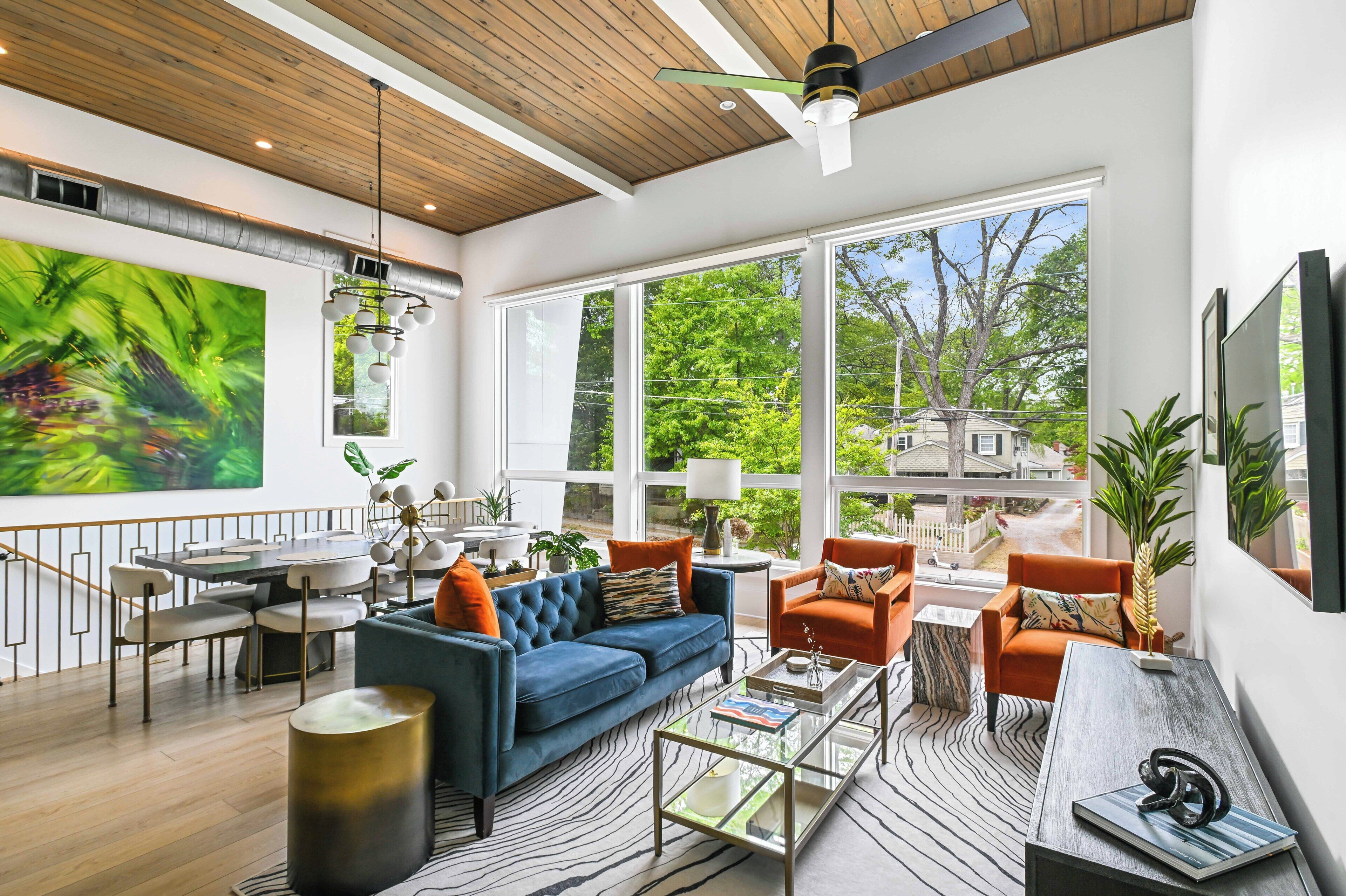 A spacious living area with a large window, a blue sofa, orange chairs, and a dining table.