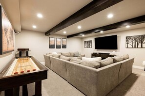 Un salon comprenant une table de jeu de «shuffleboard», un grand canapé d’angle, un téléviseur à écran plat et des œuvres d’art accrochées aux murs.