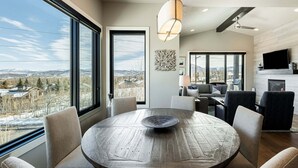 A modern living room with a round wooden table, comfortable seating, a large window with a view, a ceiling fan, and a flat-screen TV.