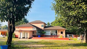 A two-story house with a large glass window on the second floor, a red roof, and a grassy yard. - 7Zee View (Nakhon Nayok)