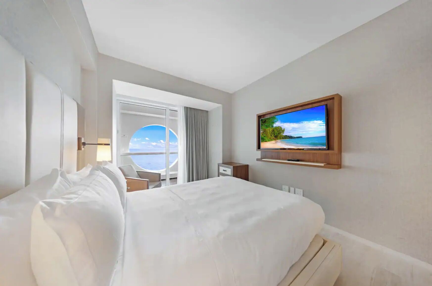 Une chambre d’hôtel avec un grand lit, un téléviseur fixé au mur, un bureau et une vue sur la mer par une fenêtre circulaire.