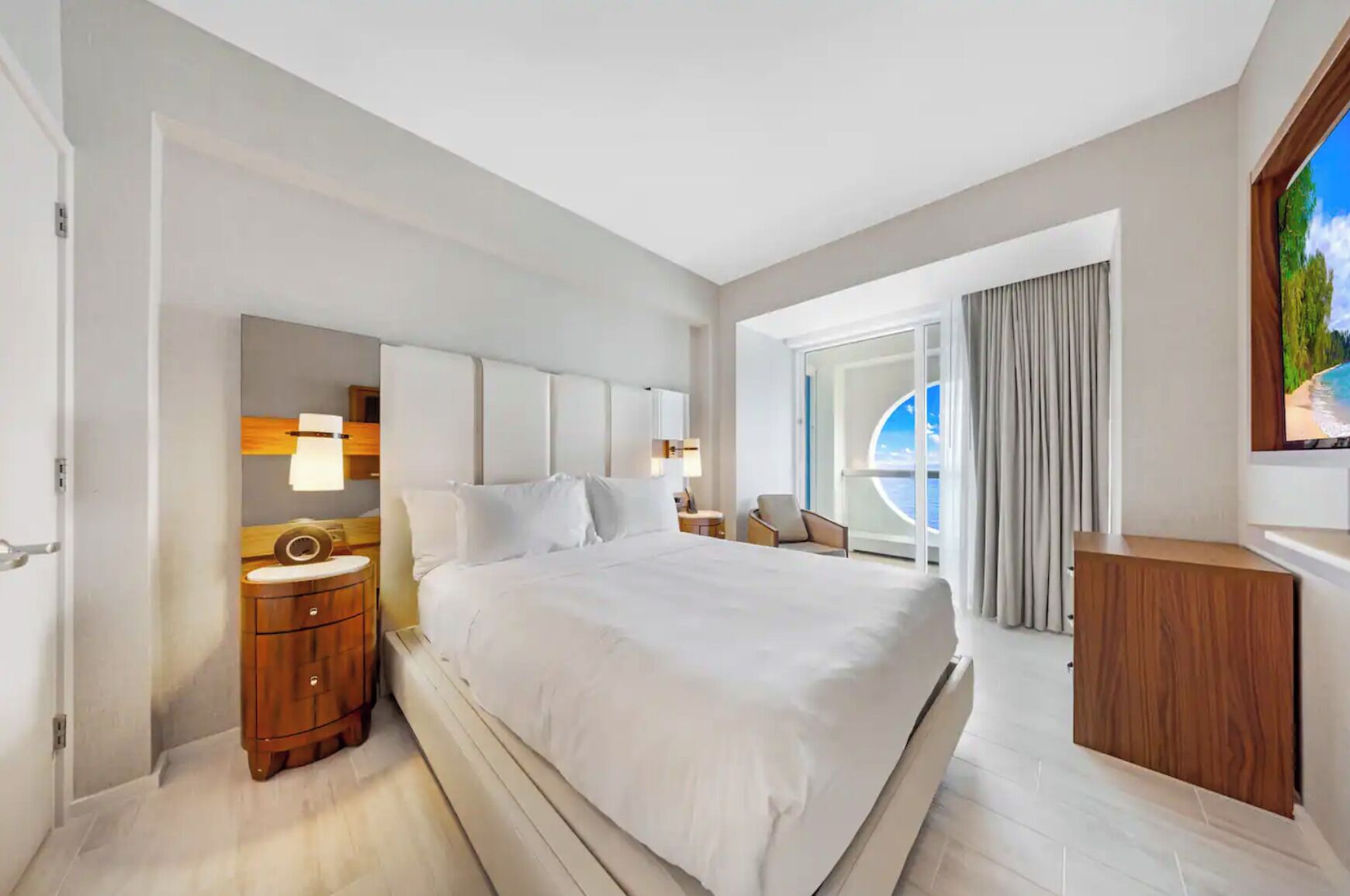A modern hotel room with a large bed, a wooden nightstand, a desk, and a view of the ocean through a large window.