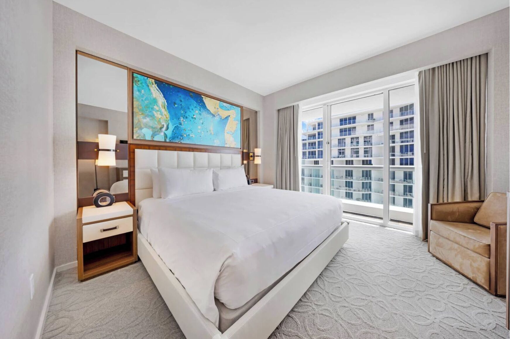 A hotel room with a large bed, a desk with a lamp, a chair, and a large window with a city view.