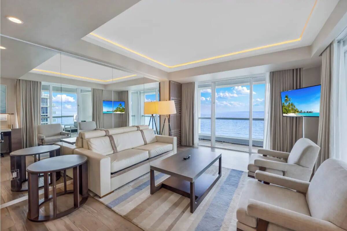 Quarto de hotel moderno com um sofá grande, uma mesa de centro, uma televisão e vista para o mar.