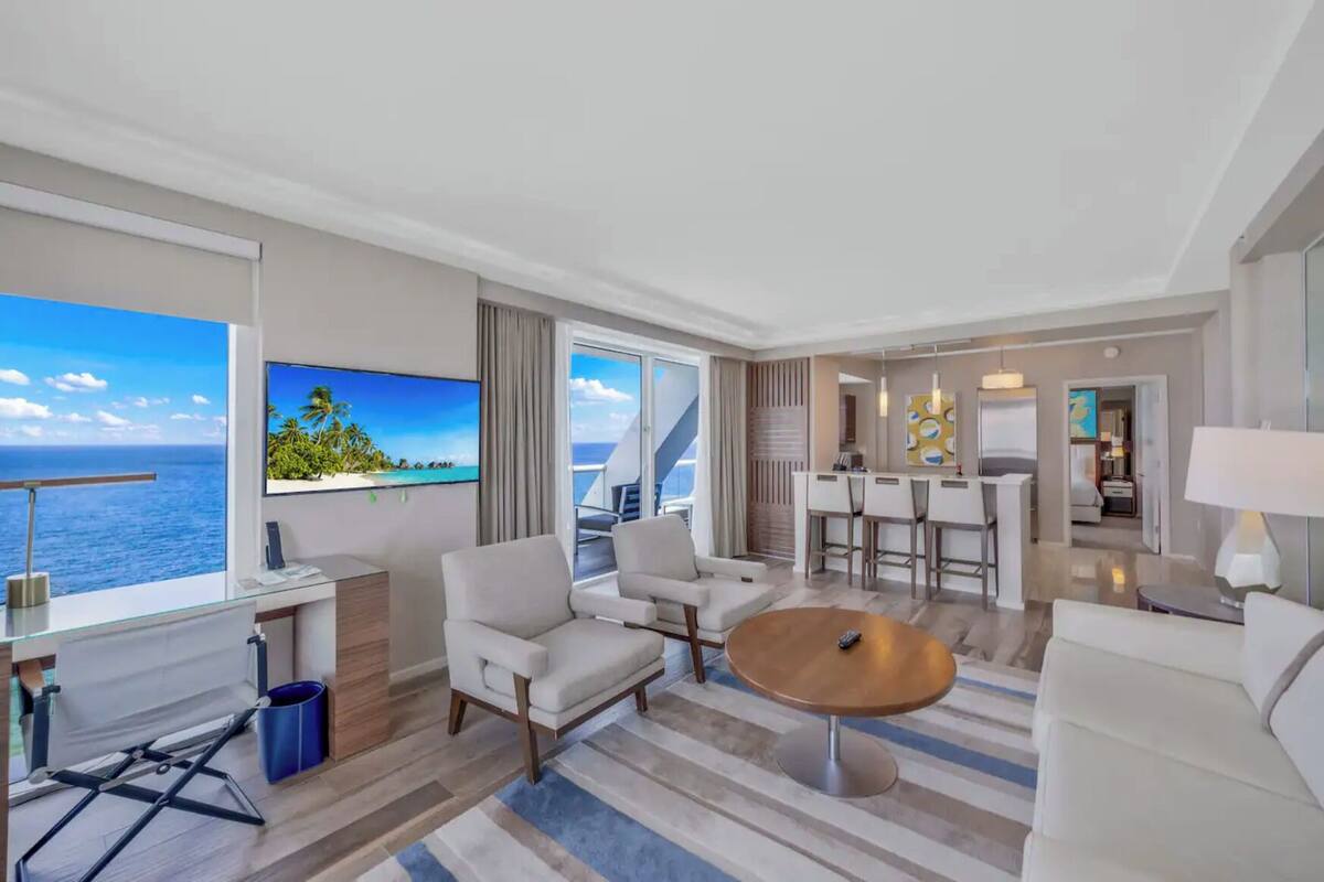 Quarto de hotel moderno com uma grande janela com vista para o mar, um espaço de estar com uma mesa de centro redonda e uma área de jantar com um bar.
