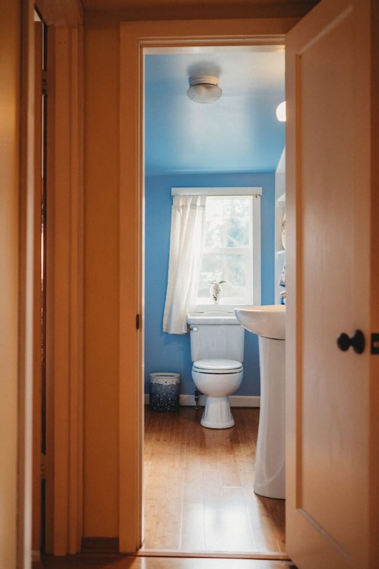 Un couloir étroit mène à une salle de bain équipée de toilettes, d’un lavabo et d’une fenêtre avec un rideau.