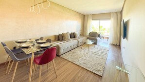 A modern living room with a dining area, a sofa, a coffee table, and a TV mounted on the wall. - Elegant Family Apartment in the Heart of Marrakech | Golf, Pool and Lake Views (Marrakech)