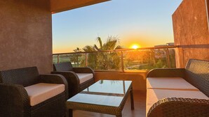 A balcony with wicker furniture, a glass table, and a view of the sunset. - Elegant Family Apartment in the Heart of Marrakech | Golf, Pool and Lake Views (Marrakech)