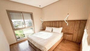 A bedroom with a large bed, wooden flooring, a window with blinds, and a bedside table. - Elegant Family Apartment in the Heart of Marrakech | Golf, Pool and Lake Views (Marrakech)