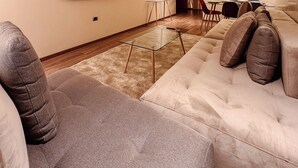 A modern living room with a large flat-screen TV mounted on the wall, a glass coffee table, a beige sofa, and a dining area in the background. - Elegant Family Apartment in the Heart of Marrakech | Golf, Pool and Lake Views (Marrakech)