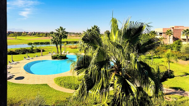 A resort with a swimming pool, palm trees, and a view of distant buildings. - Elegant Family Apartment in the Heart of Marrakech | Golf, Pool and Lake Views (Marrakech)